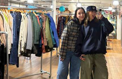 Two young women pose for a photo taken with a mobile phone in a mirror. Behind them are several racks of clothing, including coats and jackets.