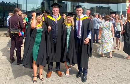 Four people in graduation gowns and caps stand together outside, smiling for a group photo. Other graduates and attendees are seen in the background. The setting is a bright, sunny day.