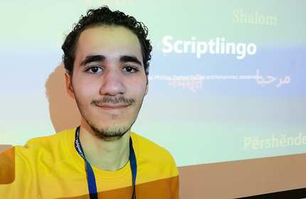 A person wearing a yellow sweatshirt and a lanyard is smiling at the camera. Behind them is a presentation screen displaying the word Scriptlingo and various greetings in different languages.