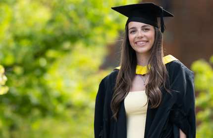 A young woman wearing a graduation cap and gown stands smiling. In the background is green foliage.
