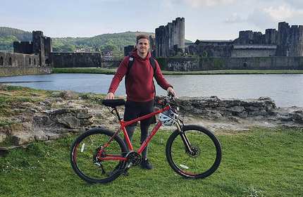 A person in a red hoodie stands next to a red bicycle on grassy terrain, with a large, historic stone castle and moat in the background under a partly cloudy sky.