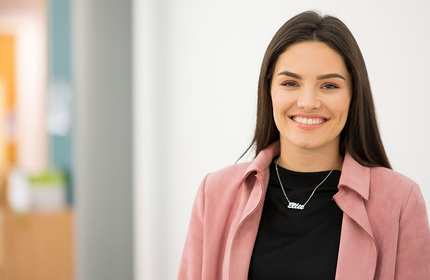 A woman with long dark hair smiles while wearing a pink jacket over a black top in a brightly lit room. She has a necklace with a name on it. The background is softly blurred, suggesting an office or indoor setting.