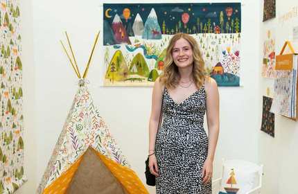 A woman in a patterned dress stands smiling next to a colorful, kid-sized teepee. The room is decorated with whimsical, illustrated landscapes on the walls and features playful designs on the wallpaper.