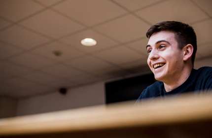 A smiling person sits in a lecture room.