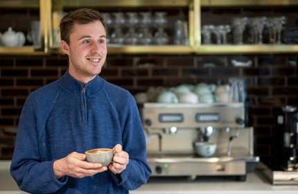 A person holding a coffee cup stands in a bar area. Behind them is a professional coffee machine.