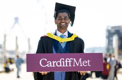Kush Shinde, in graduate cap and gown, holds a purple Cardiff Met sign with both hands