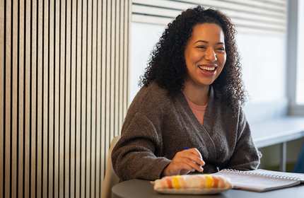 A person with curly hair sits at a table, smiling, holding a pen over an open notebook. The background features wooden paneling and sunlight streaming in through a window. A colorful pencil case lies on the table.