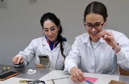 Two women in lab coats are sitting at a table, working and smiling. They have various tools and materials in front of them, including a phone, a dental mold, and colored paper.