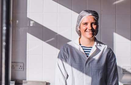 A person wearing a white lab coat and a hairnet stands smiling in a brightly lit room with white tile walls. Shadows from window panes fall across the wall, creating a pattern of light and shadow.