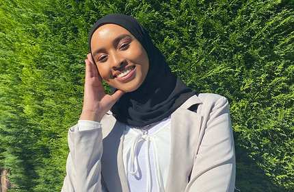 A woman wearing a hijab and light-colored jacket smiles while standing in front of a lush green hedge. She is holding her hand to the side of her face.