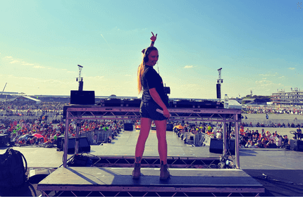 A DJ stands on an outdoor stage in front of a large, seated audience. The sky is clear and sunny. The DJ, with long hair, is making a peace sign with one hand. In the background, there are buildings and structures.