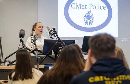 A woman is speaking at a podium with several microphones in front of her. A logo for CMet Police is displayed on a screen behind her. Several people are seated, facing the speaker.