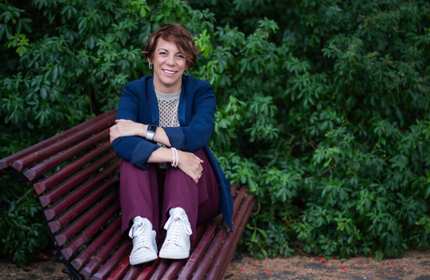 A woman sits on a bench outdoors, surrounded by lush green plants.