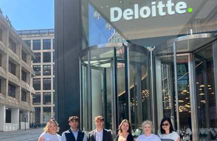 A group of young people line up outside the entrance to a building. Above the entrance's revolving doors is a sign reading Deloitte.
