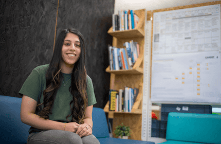 A woman with long hair sits on a blue sofa, smiling. She wears a green shirt and jeans. Behind her are bookshelves filled with books and a large poster on the wall. The setting appears to be a casual, modern office or study space.