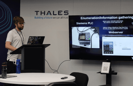 A person with a beard and glasses is standing at a podium, giving a presentation at Thales. The screen displays information on Siemens PLC and a web server. A conference room with chairs and a water bottle is visible.