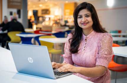 A young woman sits at a table with a laptop computer in front of her. In the background are several tables and a coffee shop area.