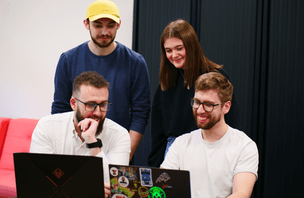 Four people are gathered around two laptops. Two people are sitting with laptops, one wearing glasses and a white shirt, the other smiling. Two people are standing, one in a yellow cap and blue shirt, the other with long hair. They appear focused and engaged.