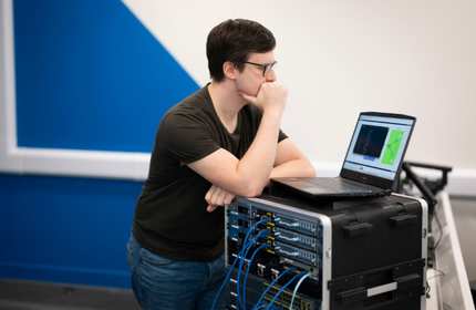 A person is focused on a laptop screen. The laptop is sitting on a rack of computer networking equipment.