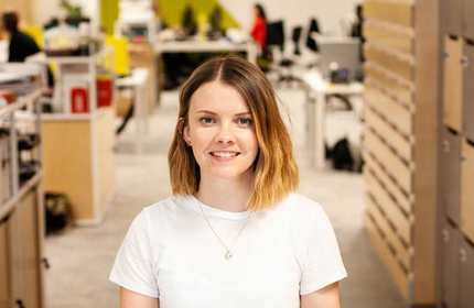 Amy Richards stands in a hallway between office cubicles and wooden storage shelves