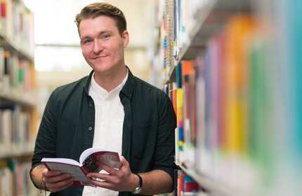 Daniel Townsend looks up from a book he's reading while leaning on a bookcase in a university library