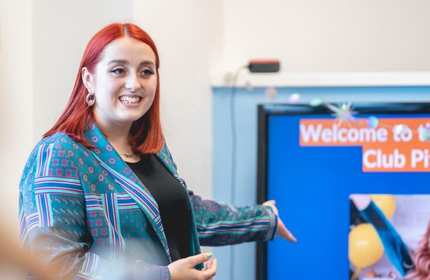Woman in a patterned turquoise blazer and black top gestures to a projector screen during a presentation