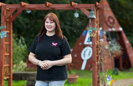 Joanne stands in front of a wooden arch and small wooden pyramid with entrance in an open garden space
