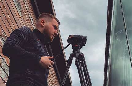 Jonathan Aveston, wearing a dark-coloured tracksuit, stands behind a tripod-mounted camcorder