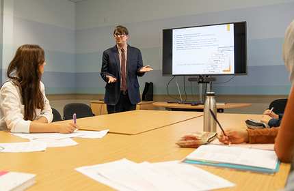 Jonathan Fry, stood on the far end of a large table, gestures with his hands during a presentation to seated attendees