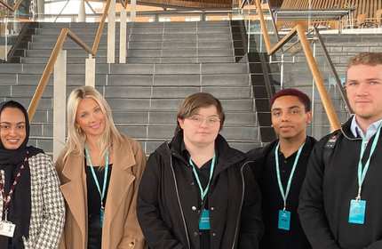A group of five young adults stand at the bottom of the stairs in front of the Senedd in Cardiff Bay