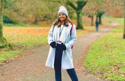Kasuni Alawattage, wearing a light blue coat and bobble hat, stands on a concrete path through a green and wooded area