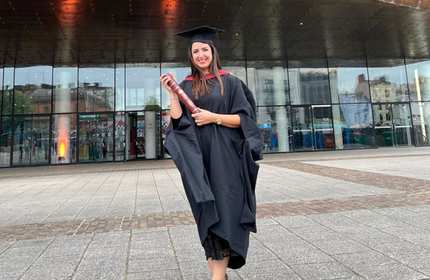 Layal Kazan, wearing a black graduate cap and gown, holds a graduation scroll tube in front of a glass-front building