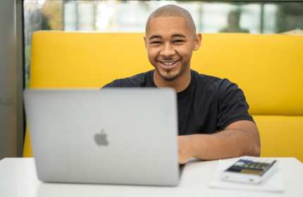 Marcus Scotland, wearing a black t-shirt, sits on a tall-backed yellow sofa while working from a laptop
