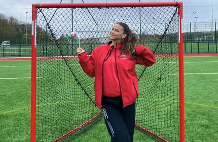 Megan Hodd, wearing a red jacket, holds a Welsh flag while standing in front of a Lacrosse net