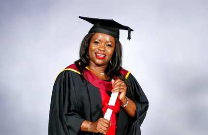 Mercy Kwaramba poses for a photograph wearing a black graduate cap and gown and holds a tied scroll with both hands