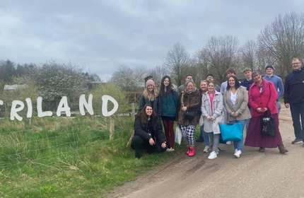 A group of adults stand beside a white sign on a grass verge