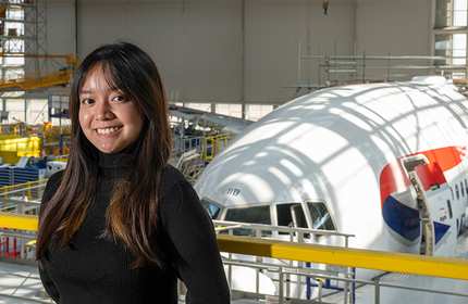 Nadia Martin, wearing a black turtleneck top, stands on a catwalk in front of an airplane in a factory hangar
