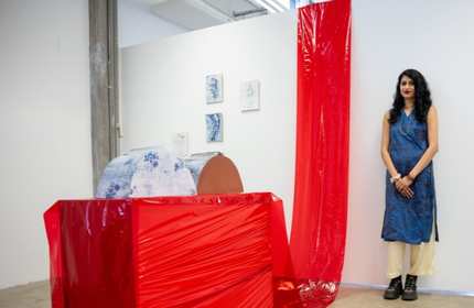 Nandini Chardavarkar stands against a white wall next to her art piece