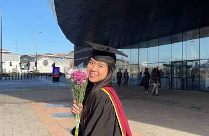 Nghi Le, wearing a graduation cap and gown, holds a small bouquet of pink and white flowers outside the Cardiff Millennium Centre