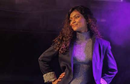 Nuwanee, wearing a textured grey turleneck and black suit jacket, poses for a photograph under purple lighting