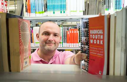 Phil Jones reaches for a book on a library shelf. He is pictured from the other side of the bookcase in a gap between the books