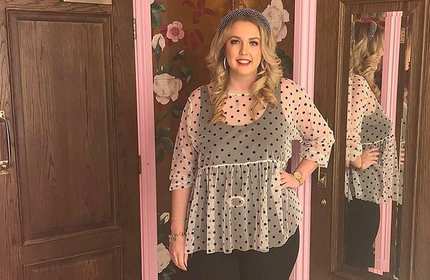 Rachel Price stands for a photograph in front of floral wallpaper