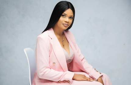 Shalom Esumeh, wearing a pink suit, sits on a white chair in front of a marble background