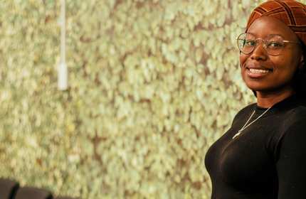Sharon Zaakura, wearing glasses and a head scarf stands in front of a textured wall