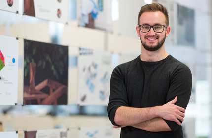 Tom Fantom stands with arms crossed in front of a glass display board containing various pictures and design illustrations