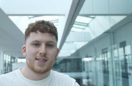 A young man with curly hair stands confidently in a well-lit hallway, showcasing a relaxed demeanor.