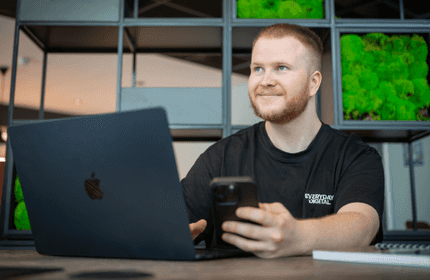 A man wearing a black shirt is seated at a table with a laptop and a cell phone in front of him.