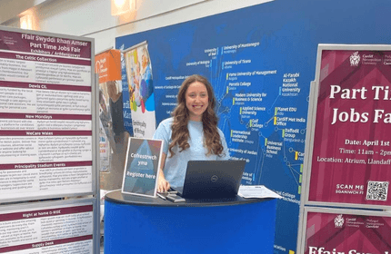 A woman stands by a table displaying a sign for a part-time jobs fair, engaging with attendees.