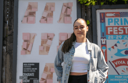 A woman stands in front of a colorful festival poster, smiling and dressed casually, with vibrant graphics in the background.