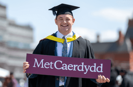 A man in graduation robes holds a sign reading 